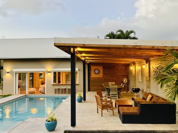 A modern outdoor pool area with a wooden pergola, seating, and a dartboard.