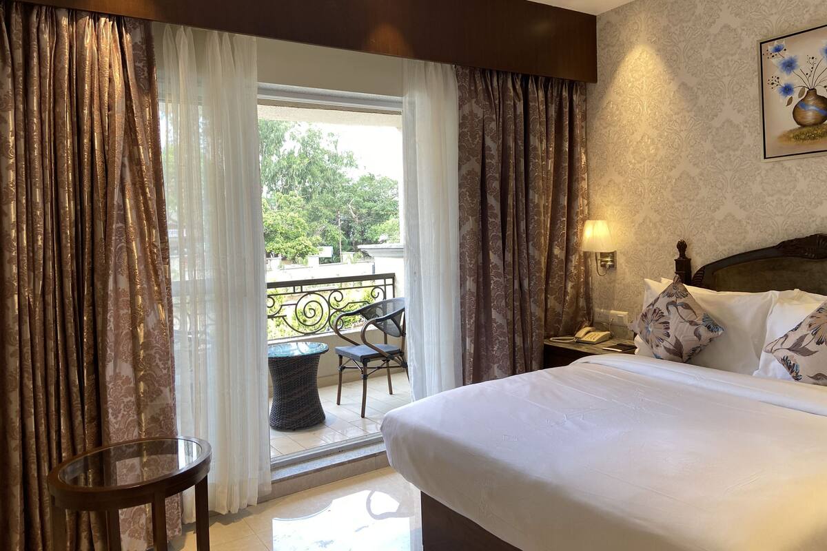 A hotel room with a bed, a nightstand, a lamp, a chair, and a balcony with a view of trees.