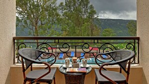 A balcony with wicker chairs, a small table with two cups and a book, and a view of a pool and mountains. - Grand Victoria The Fern Resort & Spa, Panchgani (Wai)