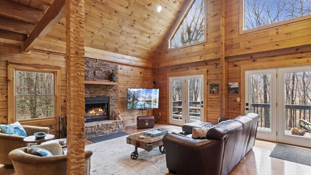 A log cabin interior with a stone fireplace, leather sofa, and large windows. - Modern Lodge | Hot Tub | EV Charger | Near Blue Ridge Pkwy (Boone)