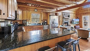 A spacious kitchen with wooden cabinets, a large island with bar stools, a refrigerator, and a stone fireplace. - Modern Lodge | Hot Tub | EV Charger | Near Blue Ridge Pkwy (Boone)