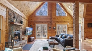 A cozy log cabin interior with a fireplace, a comfortable seating area, and a large window overlooking the trees. - Modern Lodge | Hot Tub | EV Charger | Near Blue Ridge Pkwy (Boone)