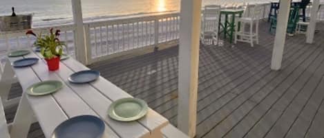 A wooden deck with a table set for a meal, overlooking the ocean at sunset.