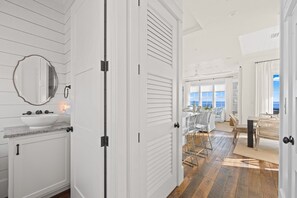 Une salle de bain moderne avec un grand miroir, un lavabo blanc et une porte donnant sur un espace de vie avec vue sur l’océan.