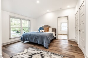 A bedroom with a large window, a bed with a blue bedspread, a patterned rug, and a door leading to another room. - La Casa Negra - Spanish Townhome in SoBro (Indianapolis)