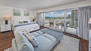Habitación de hotel con cama, sofá, mesita de noche, lámpara y vista a un puerto deportivo.