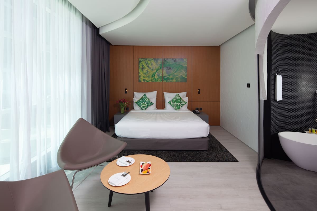 A modern hotel room with a large bed, a round dining table, a chair, a bathroom with a bathtub, and a large window with sheer curtains.