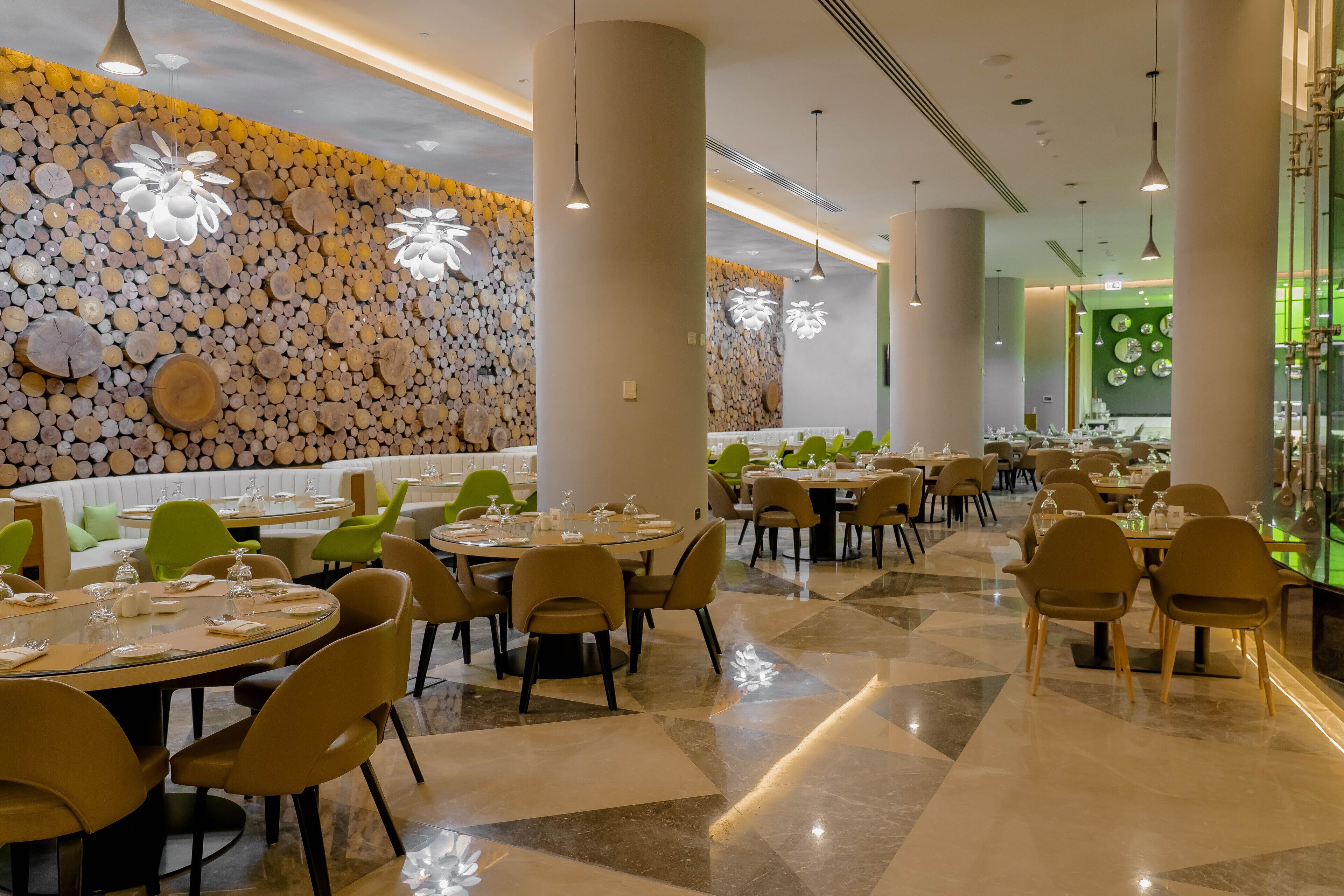 A modern restaurant with round tables, beige chairs, and a wall decorated with wooden logs.
