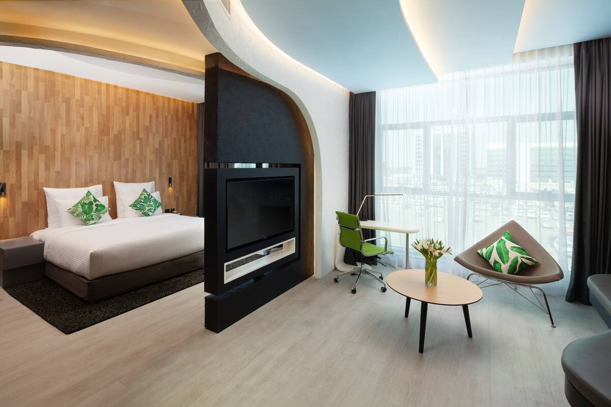 A modern hotel room with a bed, TV, seating area, and large windows.