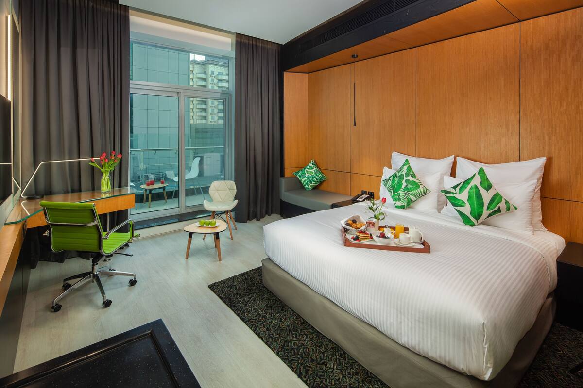 A modern hotel room with a large bed, a desk with a chair, a small table, and a window with curtains.