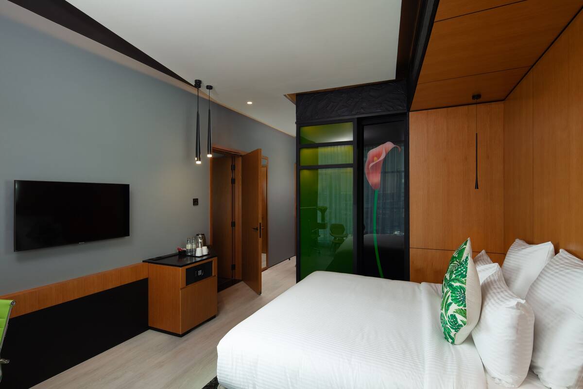 A modern hotel room with a large bed, a flat-screen TV, a wooden wardrobe, and a green glass partition.