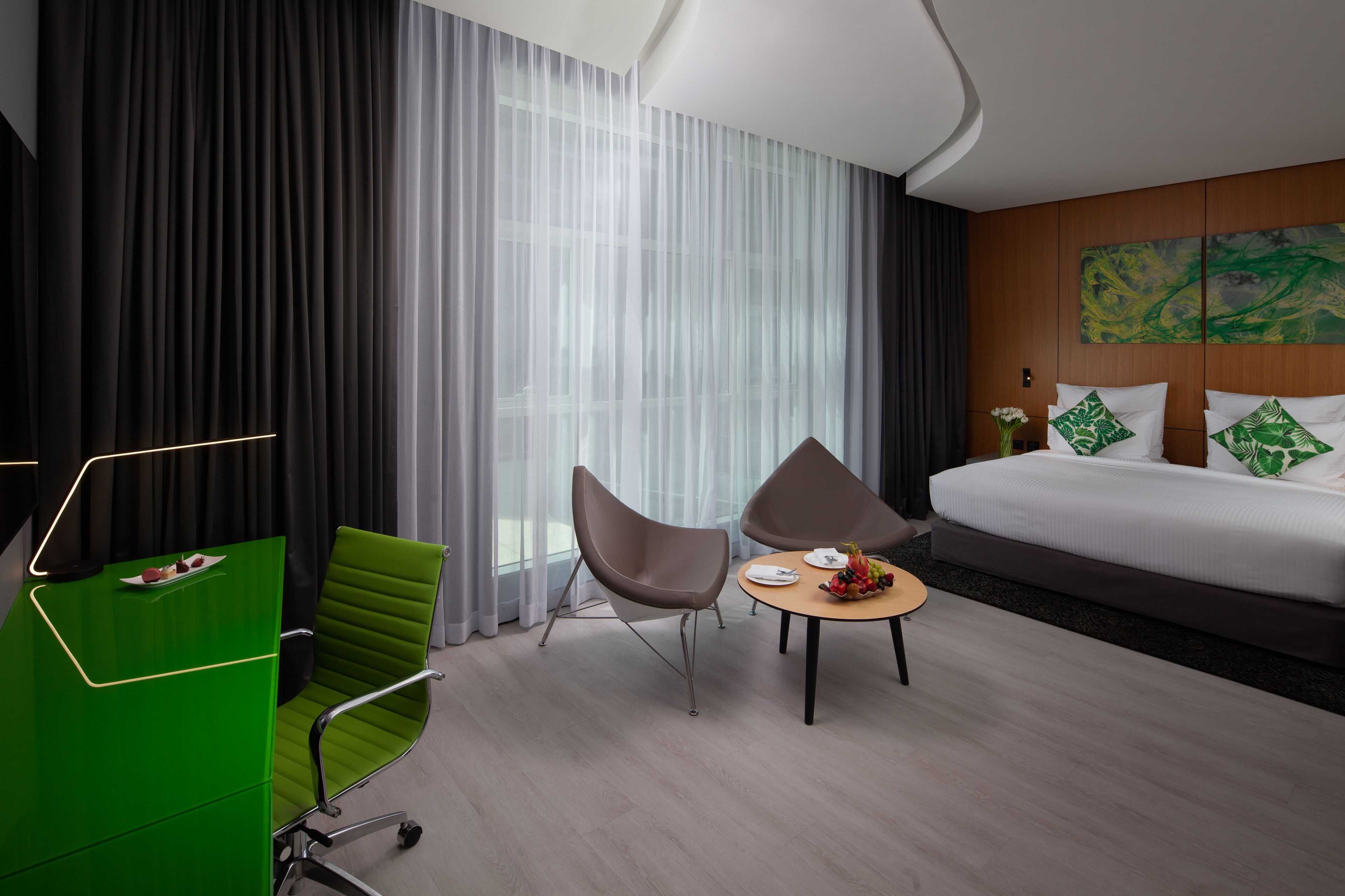 A modern hotel room with a bed, two chairs, a small table with fruits, and a desk with a laptop.