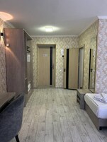 A hotel room with a wooden floor, a bed, a desk, and a chair. There are two doors, one with a 'No Entry' sign, and a mirror on the wall.