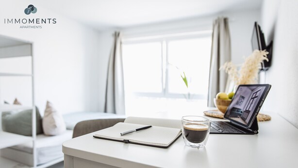 A modern, well-lit room with a white desk, a laptop, a notebook, a pen, and a glass of coffee.