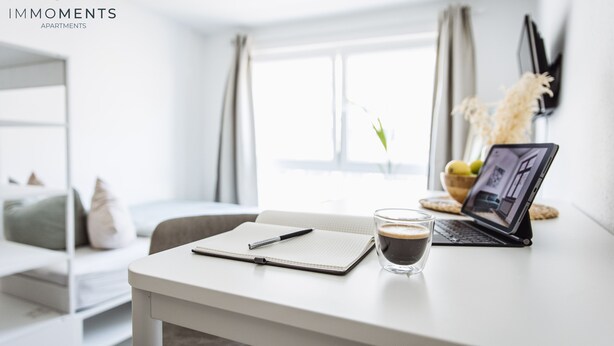 Une pièce moderne et bien éclairée, avec un bureau blanc, un ordinateur portable, un carnet, un stylo et une tasse de café.