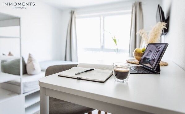 A modern, well-lit room with a white desk, a laptop, a notebook, a pen, and a glass of coffee.