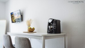 Une cuisine moderne avec une machine à café, un saladier de fruits et un vase de fleurs séchées posés sur un plan de travail blanc.