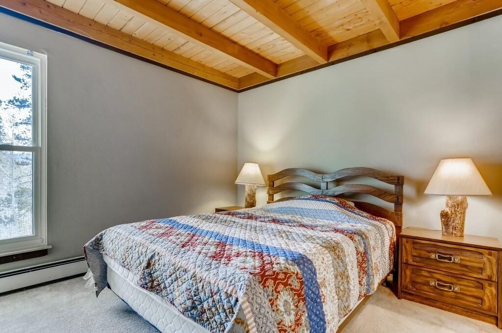 A bedroom with a wooden ceiling, a large window, a bed with a quilt, two bedside lamps, and a nightstand.