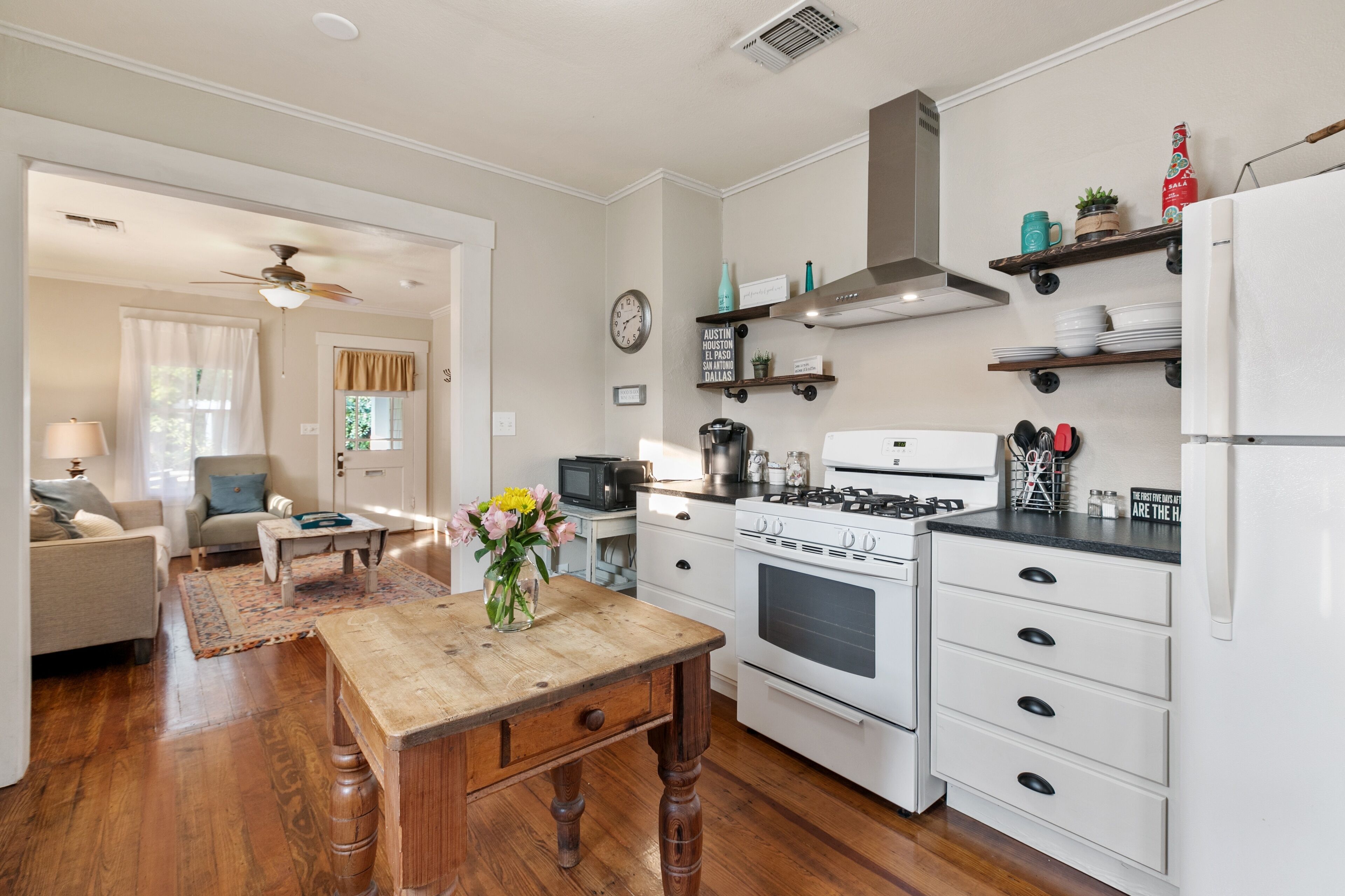 A kitchen with a wooden table, white cabinets, and a stove. There is a clock on the wall and a ceiling fan.