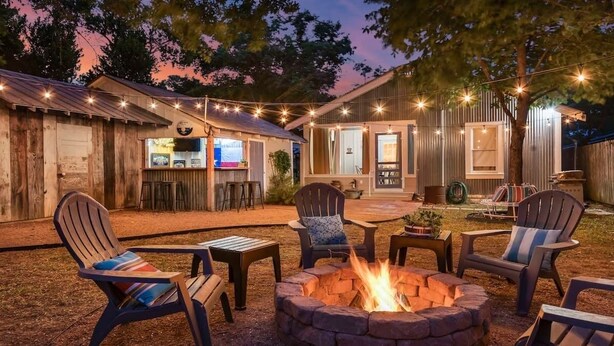 A cozy outdoor seating area with Adirondack chairs, a fire pit, and a bar area with stools and a television. - Reizend Haus 2 Bedroom Home by RedAwning (Fredericksburg)