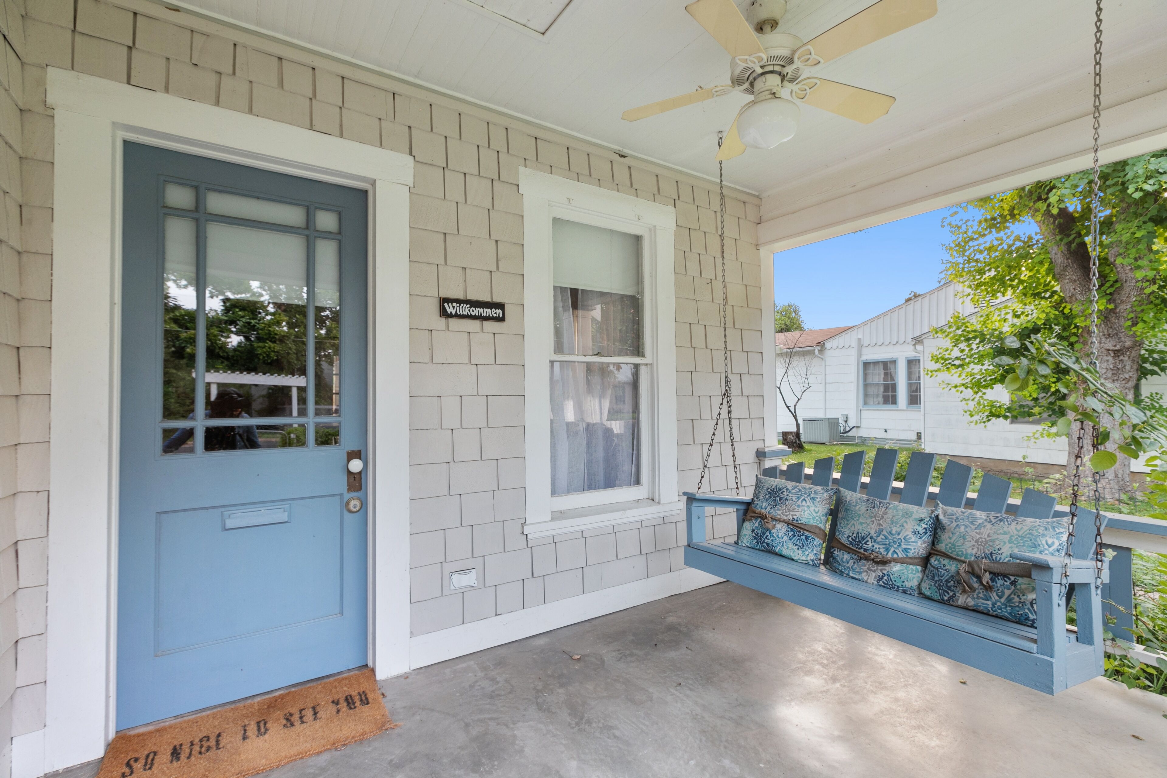 A porch with a blue door, a swing seat with patterned cushions, a ceiling fan, and a 'Welcome' sign.