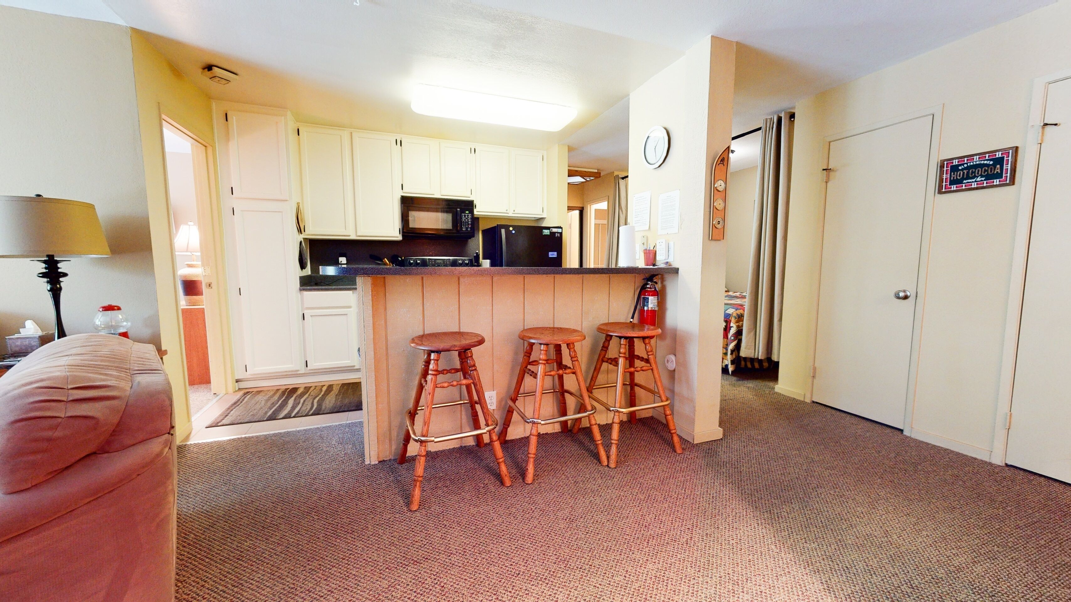 A compact kitchen with white cabinets, a microwave, and a refrigerator. There are three wooden stools at the counter.
