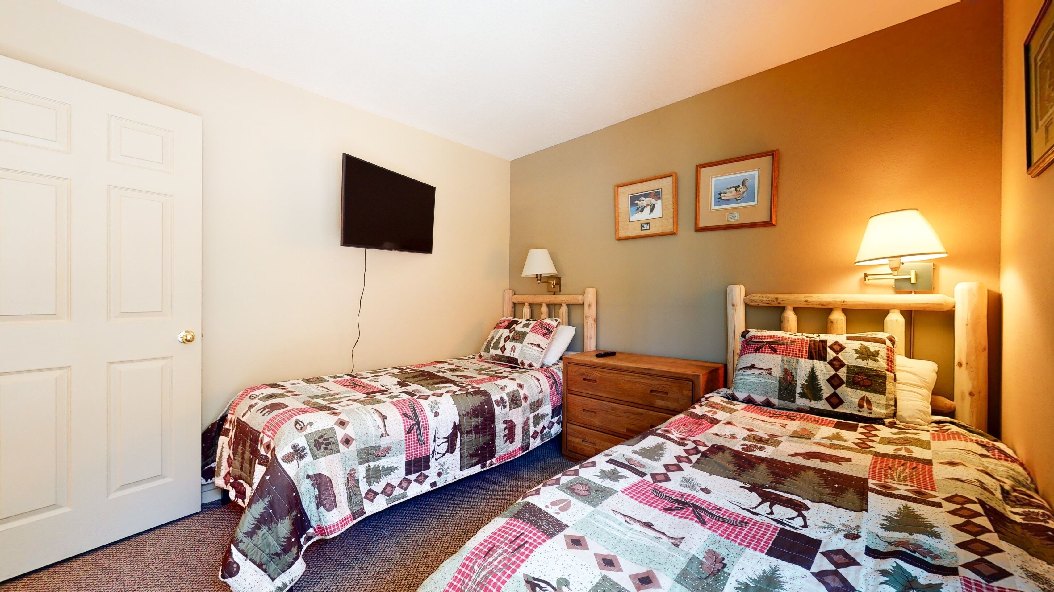 Une chambre avec deux lits simples, une commode, un téléviseur fixé au mur et une lampe sur la commode.