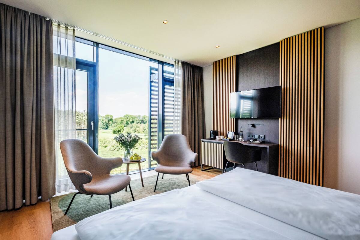 A modern hotel room with a large bed, two armchairs, a desk, and a TV.