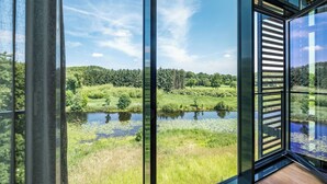 A modern interior with large glass windows offering a view of a natural landscape with a pond and greenery. - Hotel Breitenburg (Breitenburg)