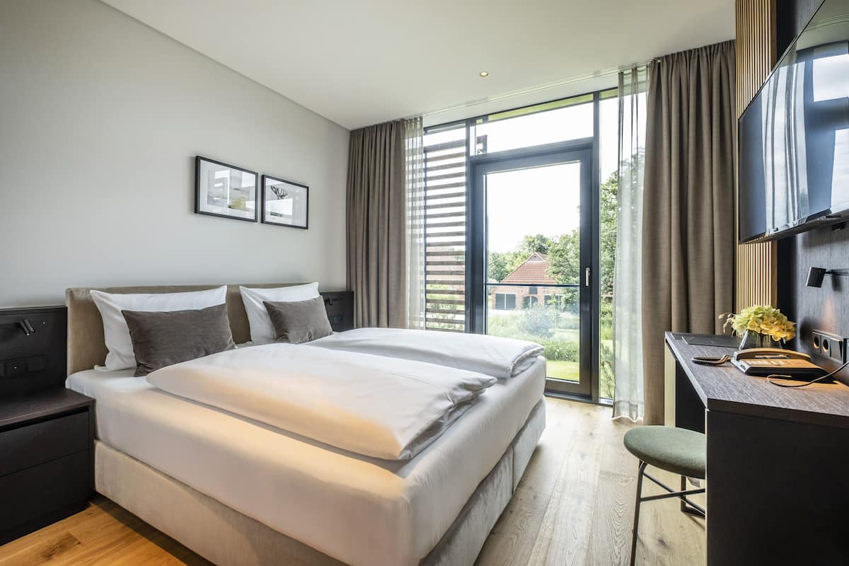 A modern hotel room with a large bed, a desk with a chair, and a view of the outdoors through a large window.