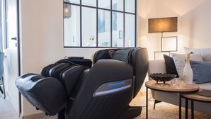 Ein Massagesessel in einem modernen Zimmer mit einem großen Fenster, einem kleinen Tisch mit einer Flasche und einer Lampe.