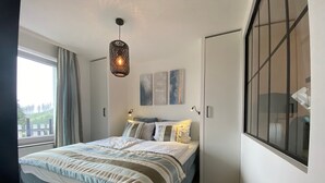 Ein Schlafzimmer mit einem großen Bett, einem Fenster mit Vorhängen, einer Hängelampe und Bildern an der Wand.