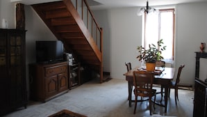 Un coin repas avec une table en bois, des chaises et une plante en pot. On aperçoit un escalier au fond.