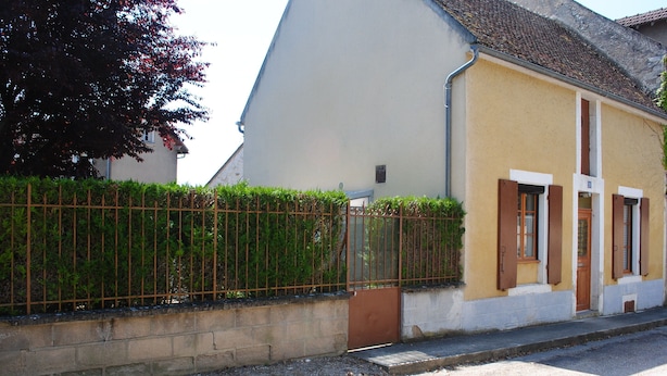 Un bâtiment à deux étages, doté d’une façade blanche et de volets bruns, d’une clôture en métal et d’un arbre aux feuilles rouges.