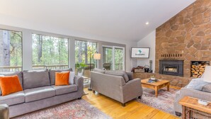 A spacious living room with a stone fireplace, large windows, and a grey sofa with orange cushions. - Near SHARC, Hot Tub, Bikes, Dog-Friendly-LEIS03 (Sunriver)