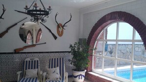A room with two striped lounge chairs, a potted plant, a chandelier, mounted rifles, and a view of a swimming pool through a large arched window.