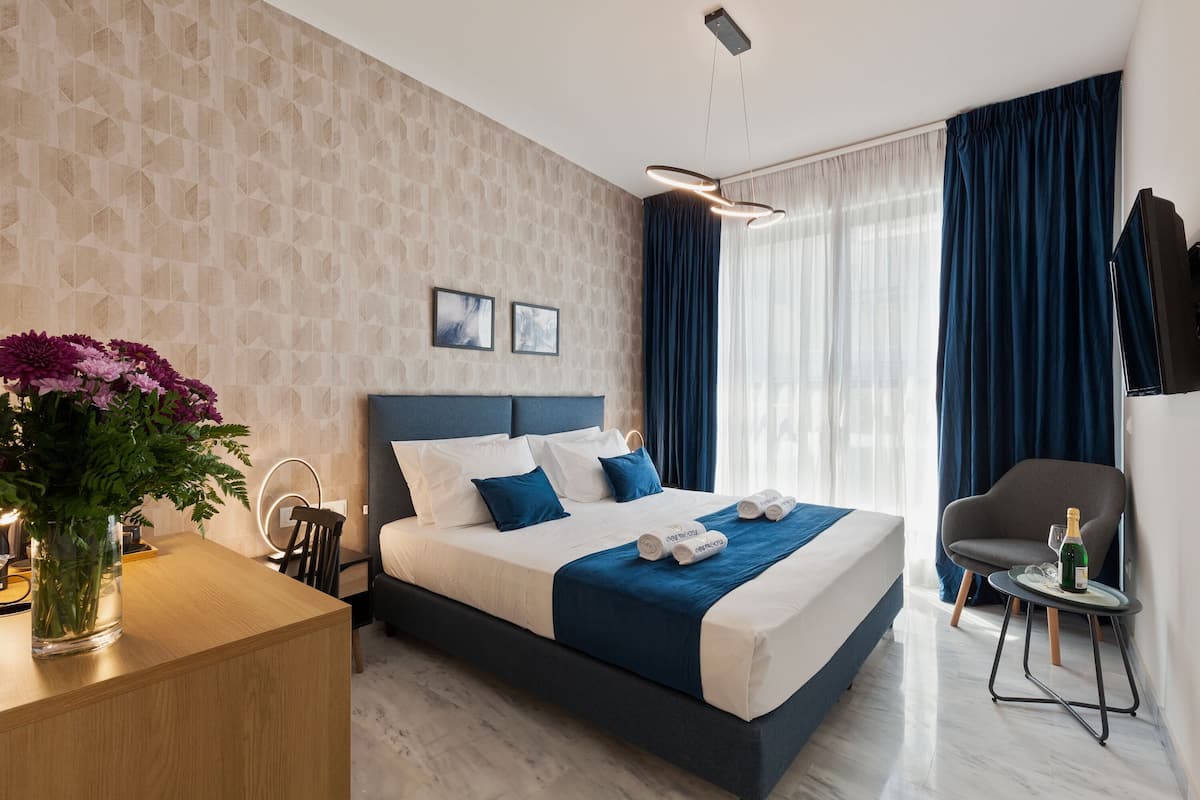 Moderni hotellihuone, jossa on suuri sänky, puinen työpöytä, tuoli, pieni pöytä, jonka päällä on pullo ja lasi, sekä maljakko kukkineen.