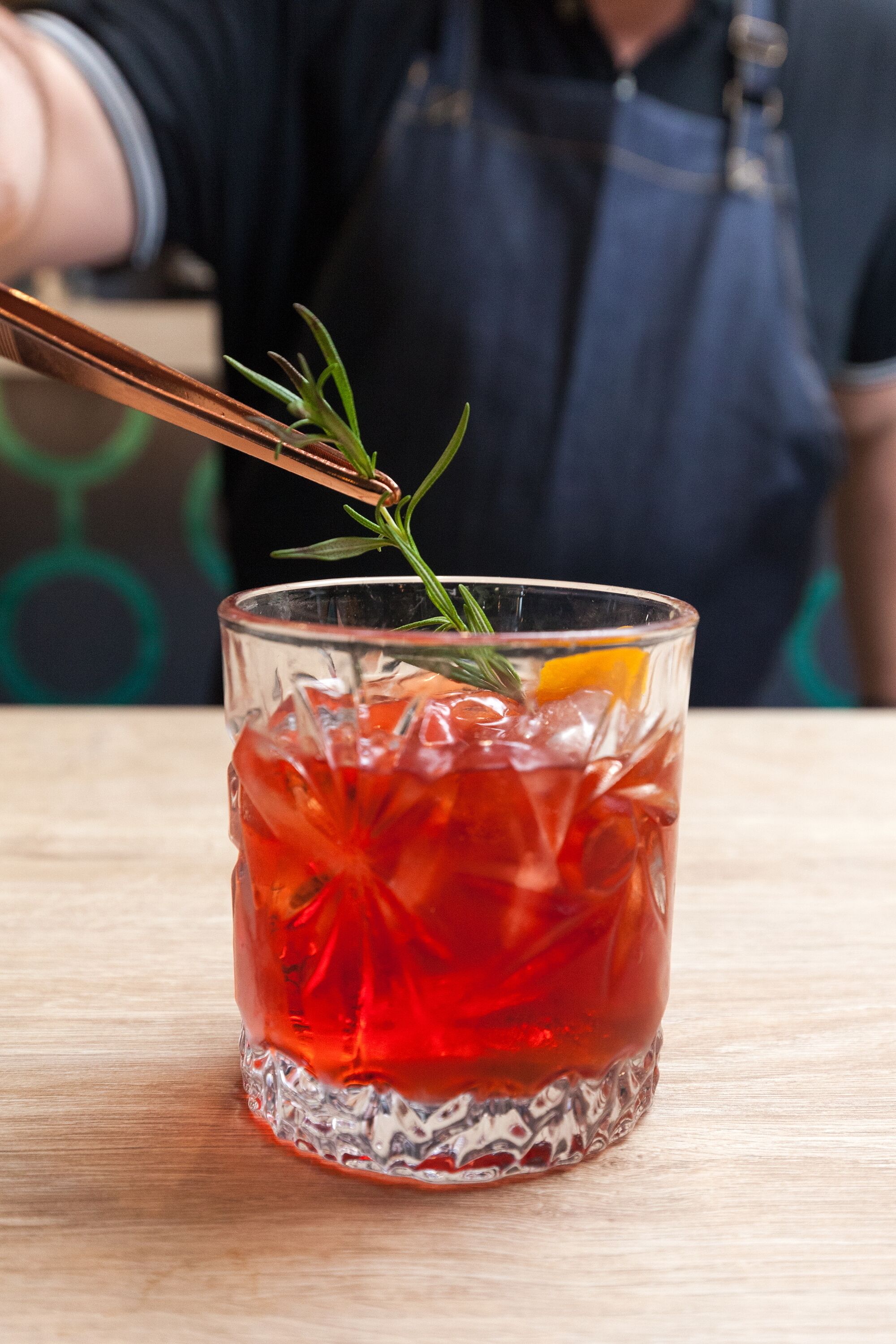 A clear glass with a red beverage, garnished with a sprig of rosemary and an orange slice, on a wooden surface.