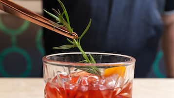 A clear glass with a red beverage, garnished with a sprig of rosemary and an orange slice, on a wooden surface.