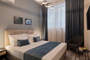 A hotel room with a bed, a chair, a small table, and a wall-mounted air conditioner.