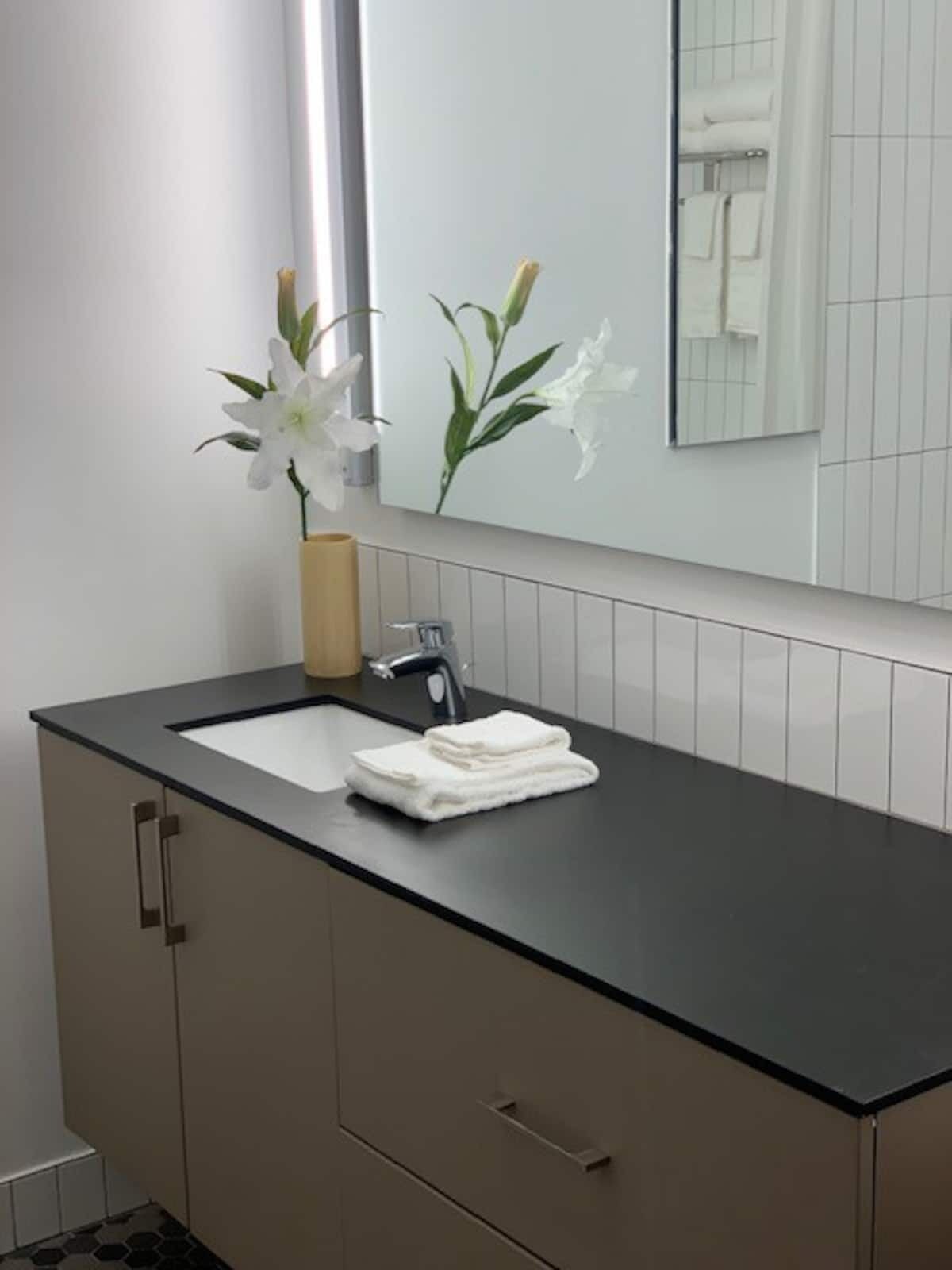 Une salle de bain moderne avec un plan de toilette noir, deux vasques, un miroir et un vase contenant des fleurs blanches.