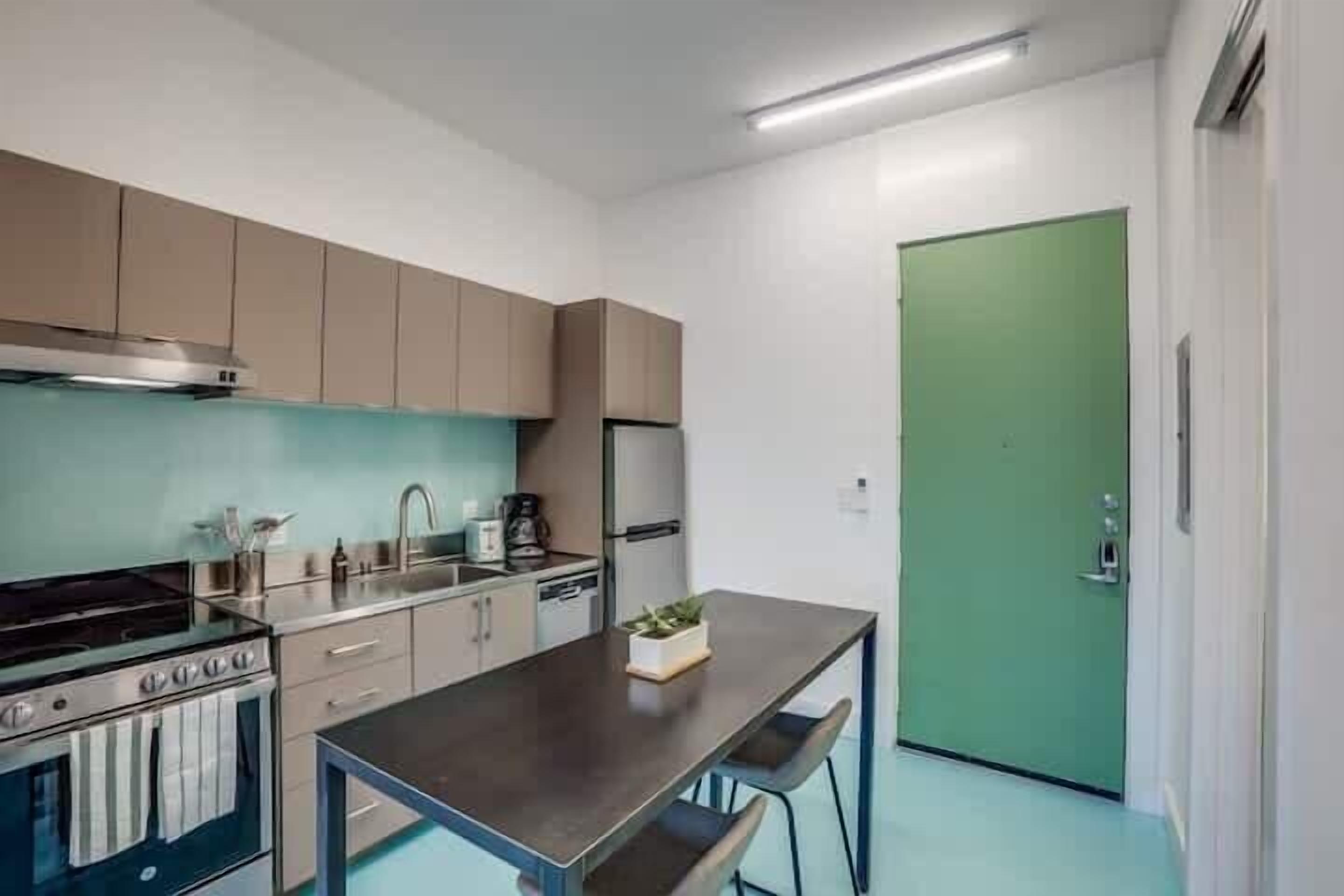 Eine kleine Küche mit Herd, Spüle und Kühlschrank, einer grünen Tür und einem Tisch mit Stühlen.