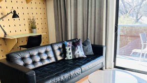 Een moderne woonkamer met een zwarte leren bank, een ronde glazen salontafel, een houten bureau met planken en een groot raam met gordijnen.