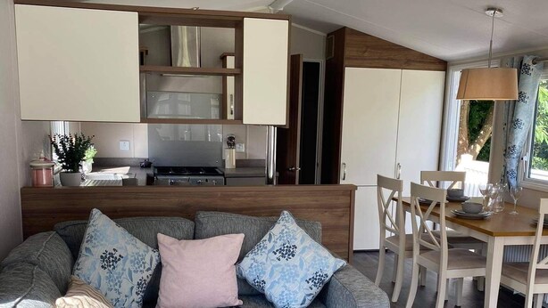 Un salon moderne avec un canapé d’angle gris, une table à manger en bois et un coin cuisine doté de meubles blancs et d’appareils électroménagers en acier inoxydable.