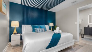 A modern bedroom with a white bed, blue accent pillows, and a teal throw blanket. There are two bedside tables with lamps and a small clock. The room has a blue accent wall and a wooden ceiling. - Disney-Themed Escape – Frozen & Star Wars Rooms, Pool, Games & Family Fun (Kissimmee)