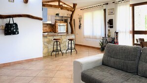 Una sala de estar acogedora con sofá, una zona de cocina con salpicadero de piedra y techo con vigas de madera.