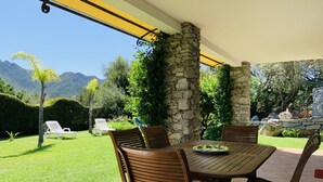 Un patio cubierto con muro de piedra, sillas de madera, una mesa con un plato de limones y vistas a un jardín exuberante y montañas.