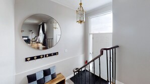A hallway with a round mirror, a chandelier, a wooden bench, and a staircase with a black railing. - CHARMING THREE BEDROOM FLAT (brampton)