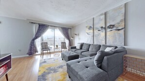 A modern living room with a grey sectional sofa, a small round table, and a chair with patterned cushions. There is a large window with purple curtains, and a painting on the wall. - CHARMING THREE BEDROOM FLAT (brampton)