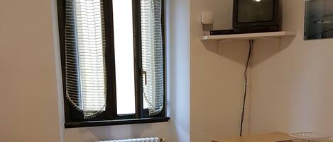 Un salon comprenant un canapé, une petite table, un radiateur, une fenêtre avec des stores et un téléviseur posé sur une étagère.